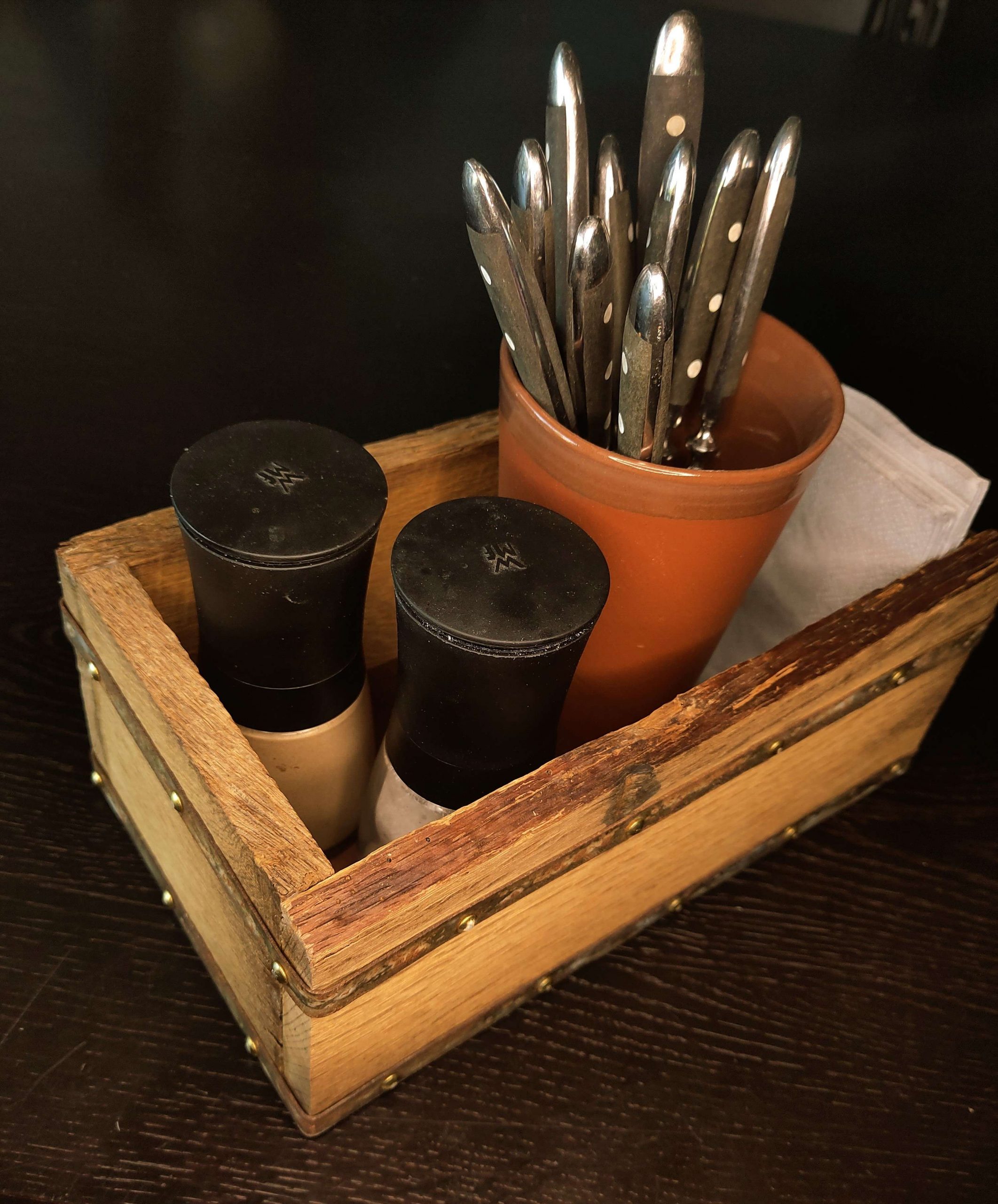 Ein Tonbecher mit Besteck, Servietten, Salz- und ein Pfefferstreuer sind im hauseigenen, rustikal charmanten Holz-Kästchen des Sorglosen Kobolds in Ludwigsburg angeordnet.
